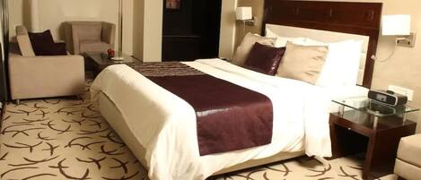 A hotel room with a large bed, a sofa, a desk with a glass top, and a lamp.