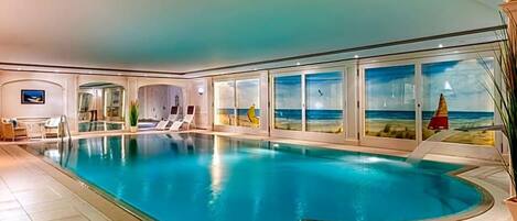 Une grande piscine intérieure aux parois en verre transparent, avec des chaises longues et une vue sur la mer.
