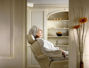 A person reclining in a chair with a white robe, a shelf with decorative items in the background, and a vase with flowers to the right. - Carstens Landhaus (Timmendorfer Strand)