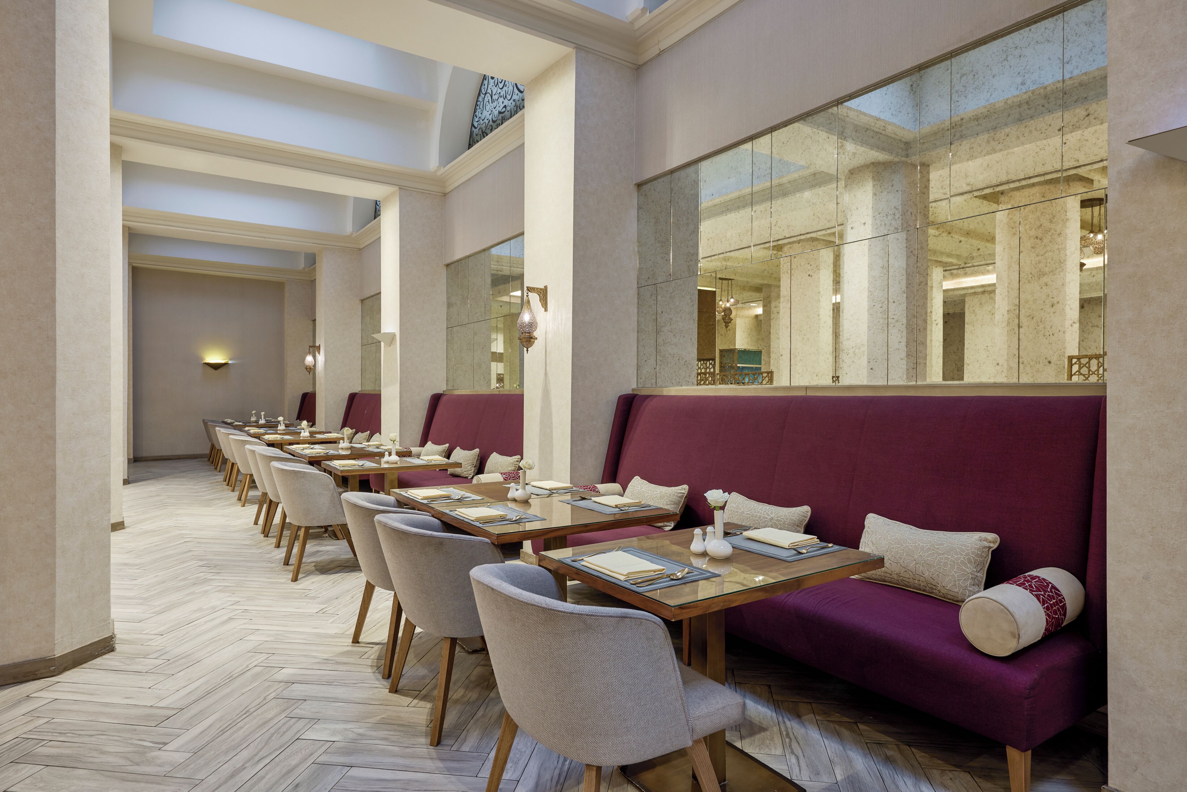 A hotel dining area with multiple tables and chairs, a large mirror, and a high ceiling.
