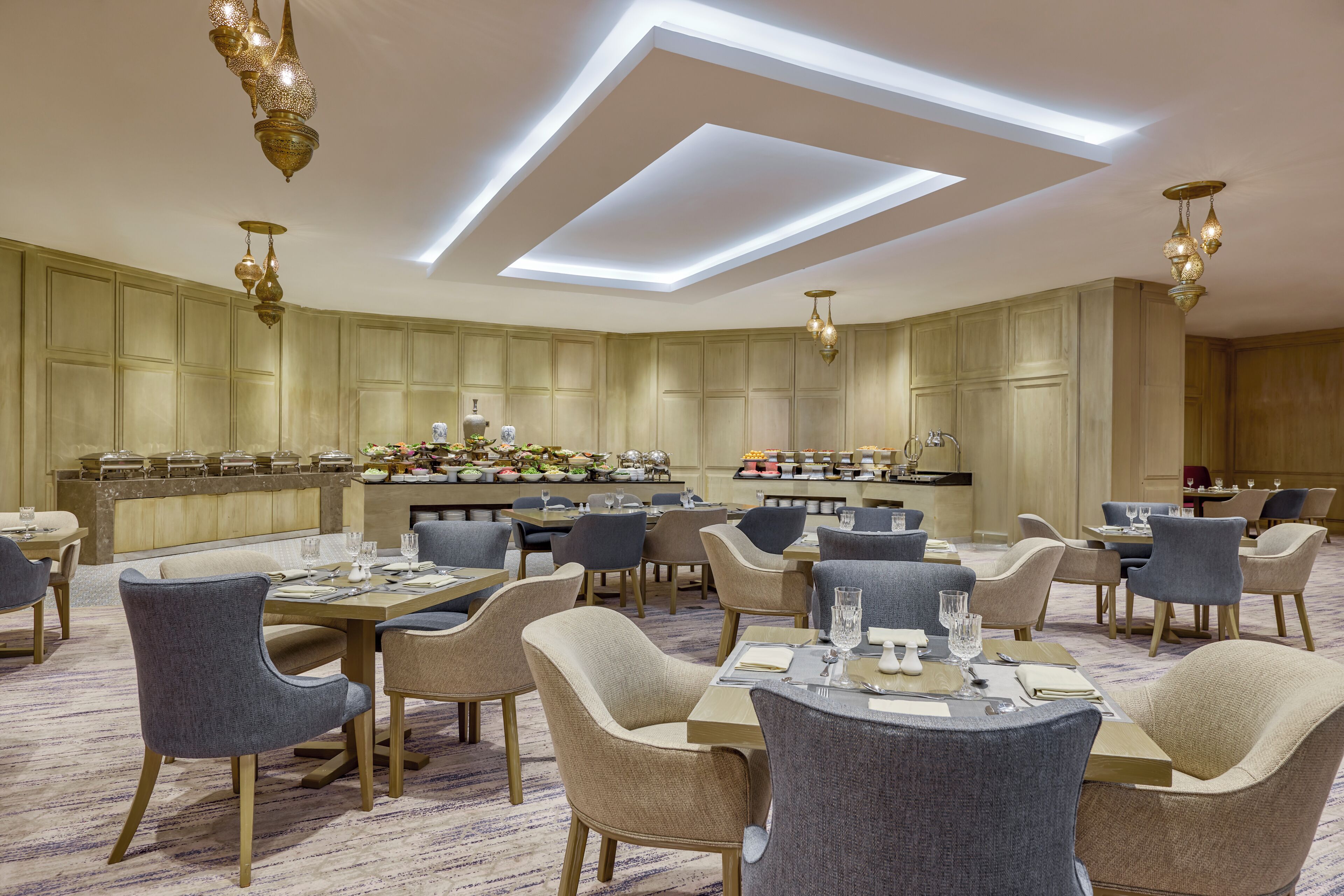 Ein geräumiger Essbereich mit mehreren Tischen und Stühlen, einBuffet mit verschiedenen Speisen sowie ein modernes Deckenleuchtendes Design mit Einbauten.