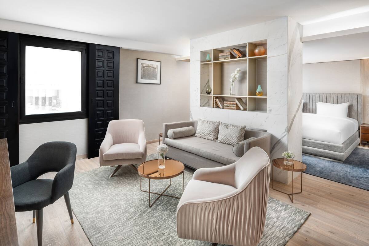 Een moderne hotelkamer met een slaapbank, twee fauteuils, een klein bijzettafeltje, een nachtkastje, een boekenkast en een bed.
