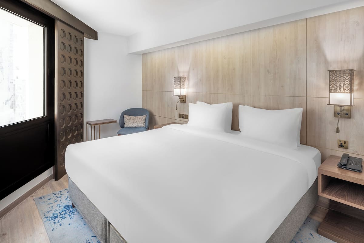 Een groot bed met witte lakens, een houten hoofdbord, twee wandlampen, een nachtkastje met een telefoon, en een raam met uitzicht op gebouwen.
