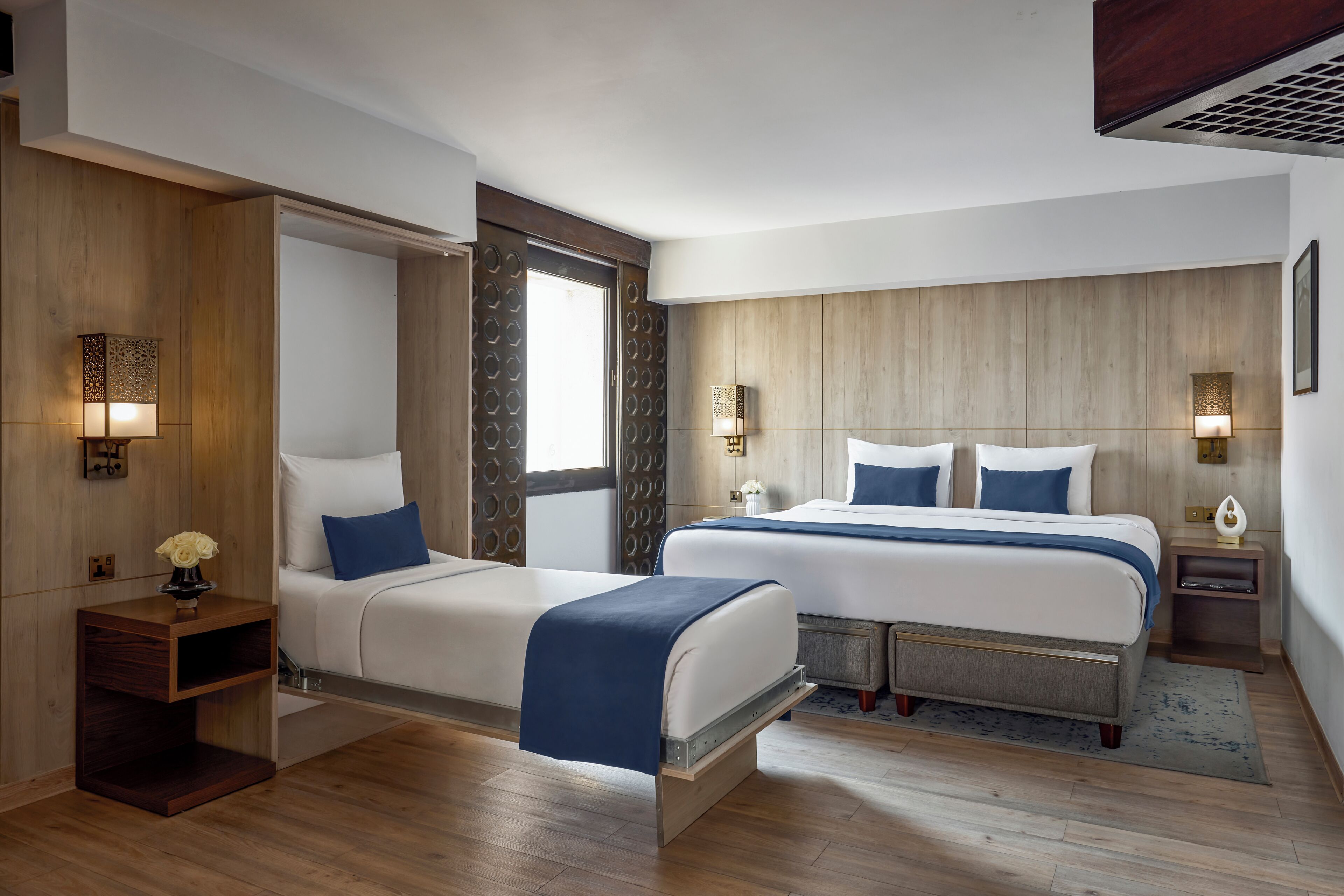 Una habitación de hotel moderna con dos camas, un armario de madera, una mesita de noche con lámpara y un ventanal grande.