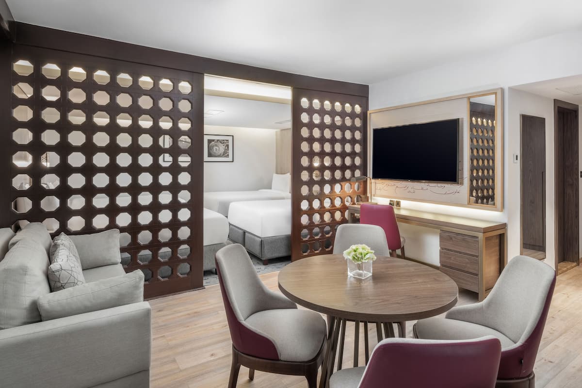 Een moderne hotelkamer met een ronde eettafel, grijze en rode stoelen, een bank, een televisie en een houten scheidingswand met ronde uitsparingen.