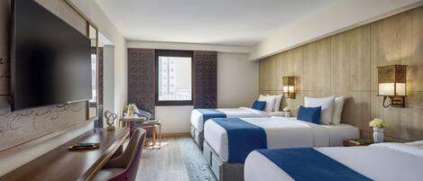 A hotel room with a flat-screen TV, two beds with blue bedding, a desk with a chair, and a large window.