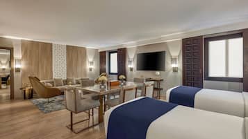 Una habitación de hotel moderna con zona de comedor, sofá, televisor y un ventanal grande.