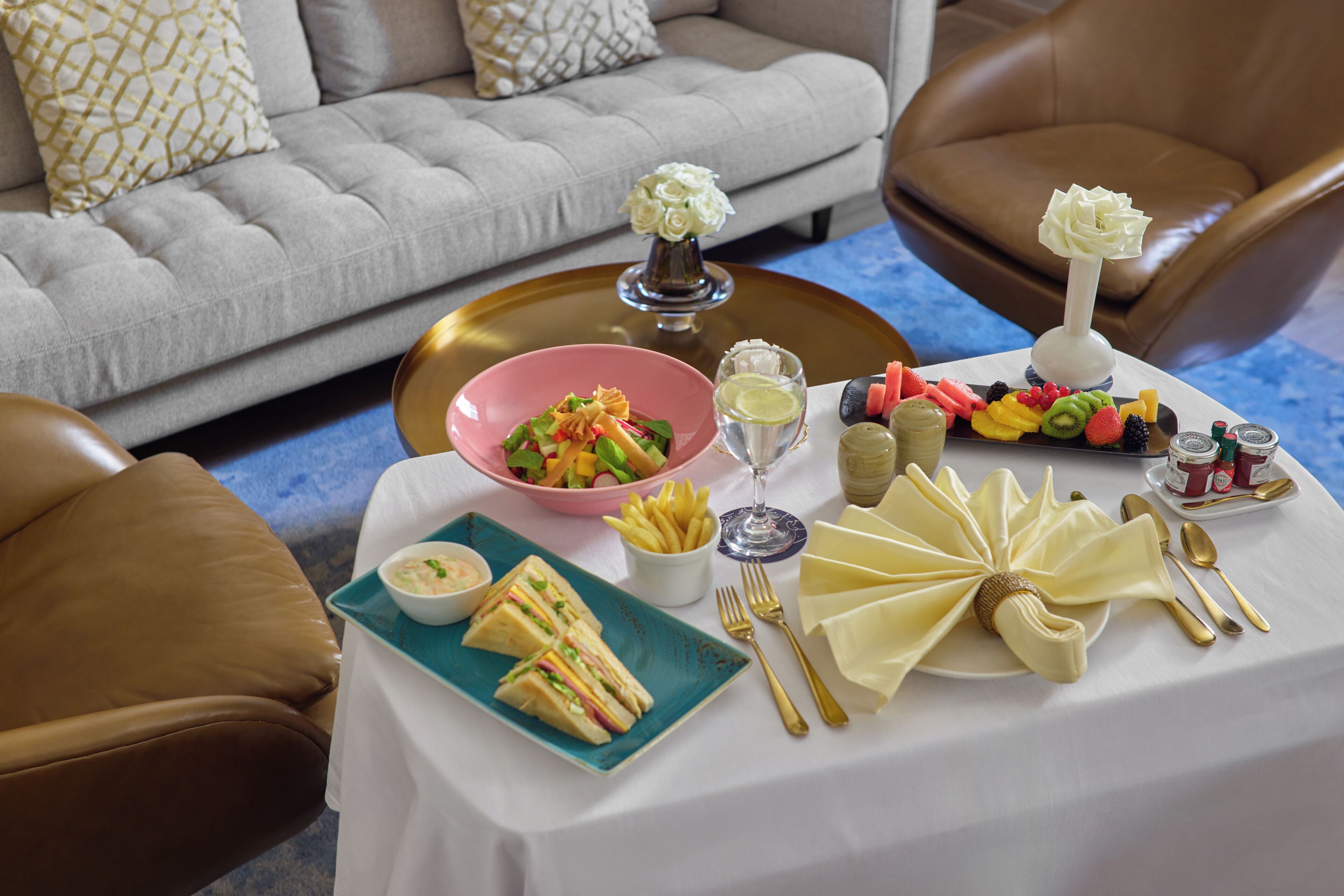 A dining table set with a variety of food items, including sandwiches, fruits, and condiments, in a room with a sofa and chairs.