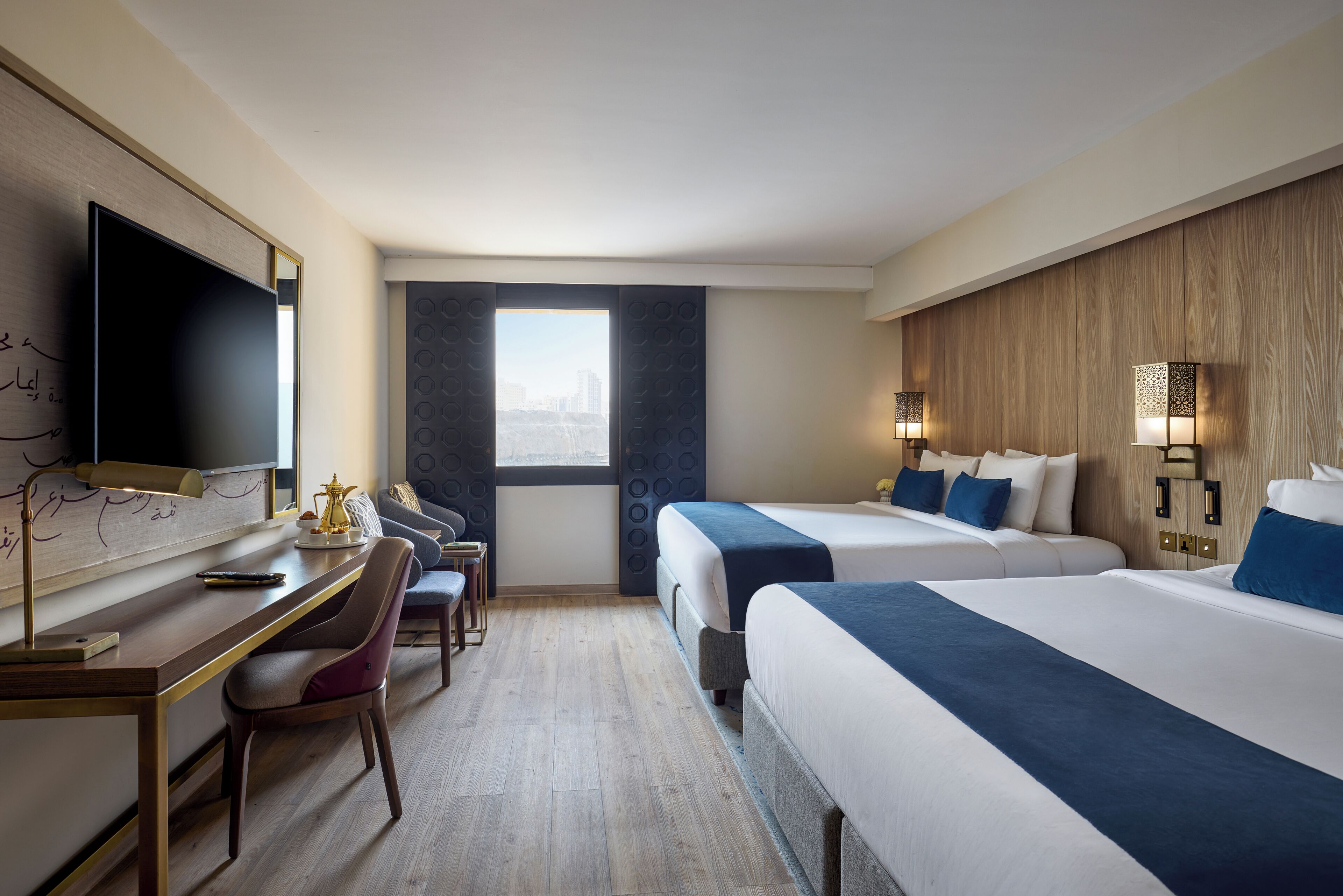 Habitación de hotel con dos camas, un escritorio, una silla, un televisor y una ventana con vistas a edificios.