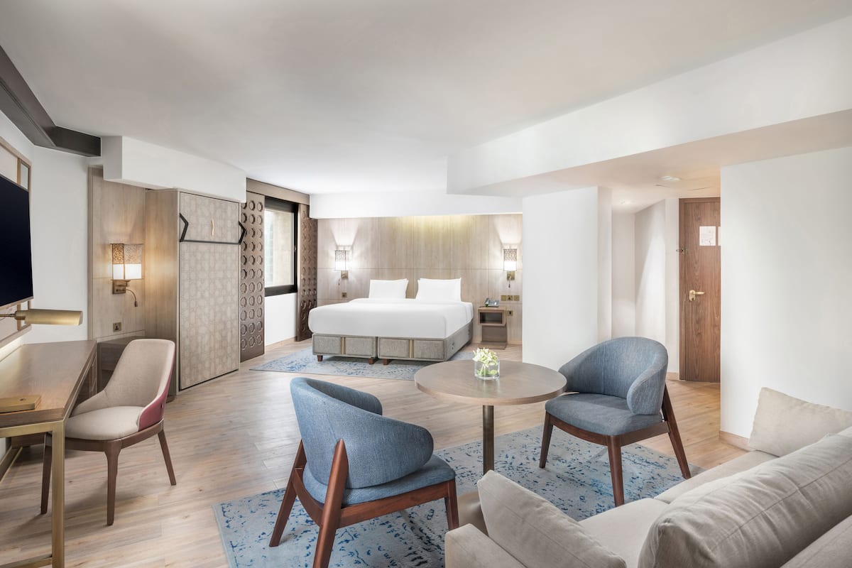 Een moderne hotelkamer met een bed, twee fauteuils, een salontafel, een bureau en een televisie.