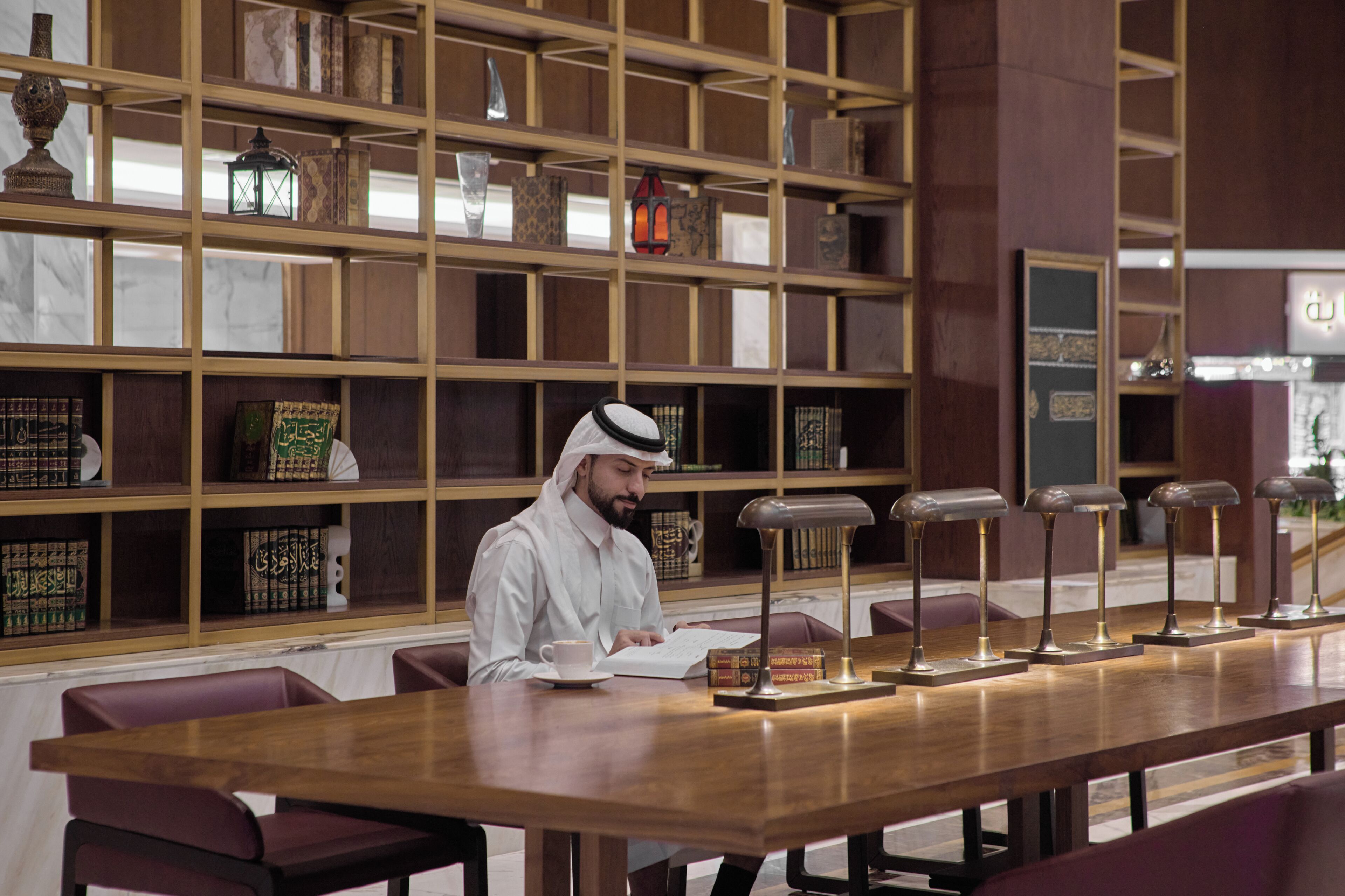 A man seated at a long wooden table in a library or reading room with bookshelves in the background.
