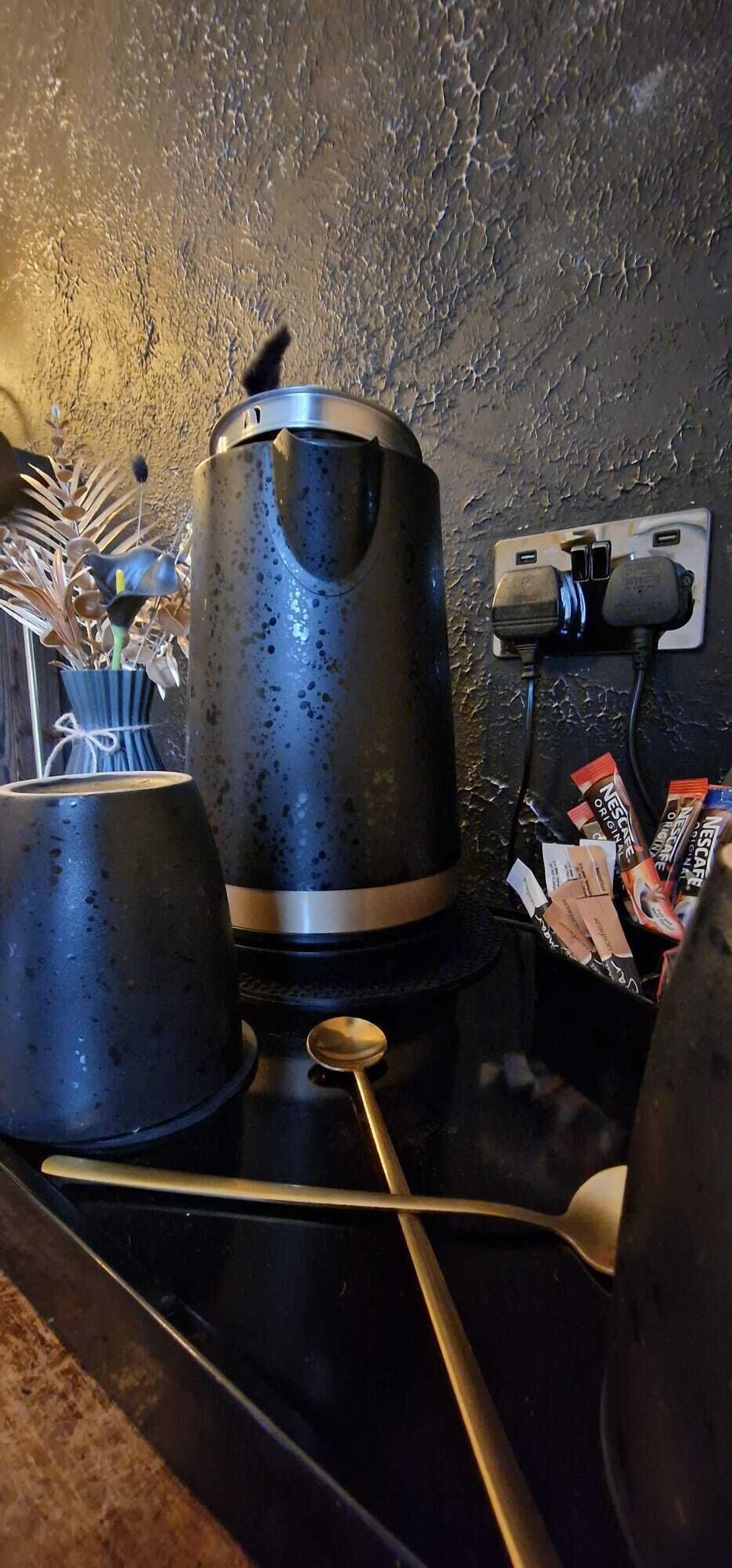 Una zona de cocina con una tetera, una taza y una cuchara sobre una encimera. Hay un enchufe y algunos embalajes en la pared.