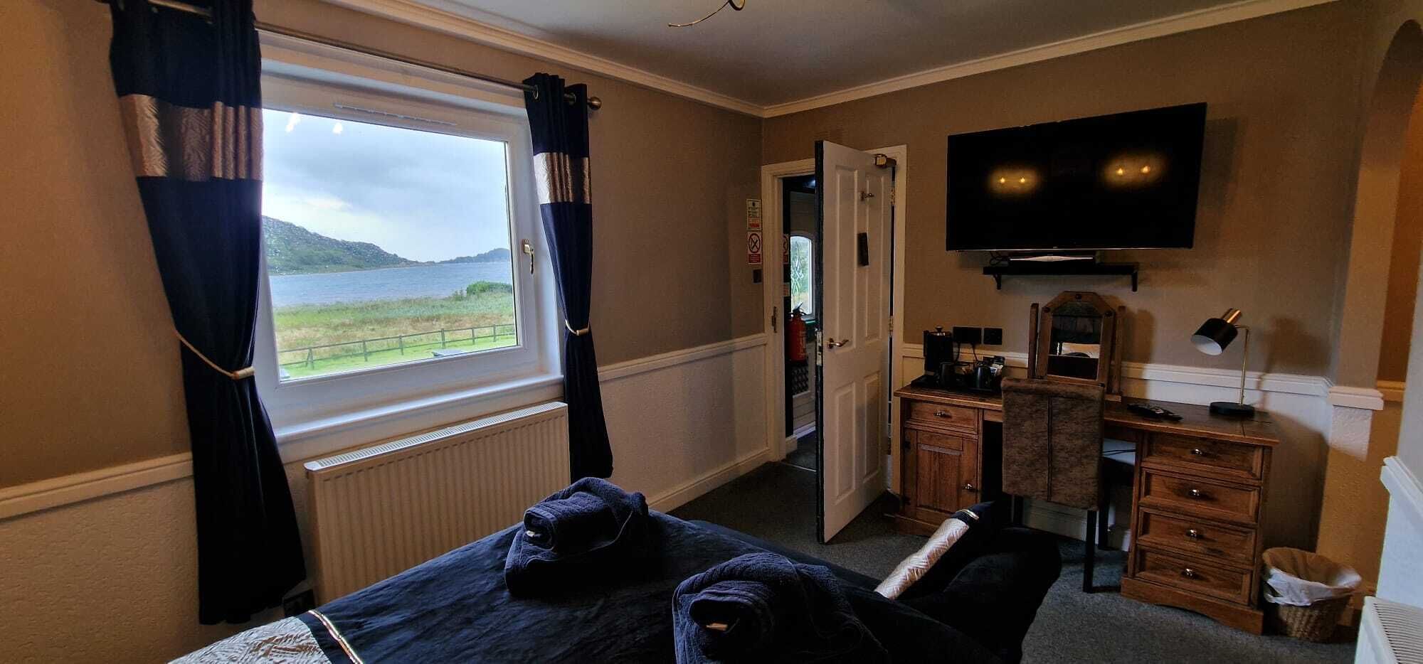 Ein Hotelzimmer mit Bett, Schreibtisch, Sessel, Fernseher und Fenster mit Blick auf Meer und Hügel.