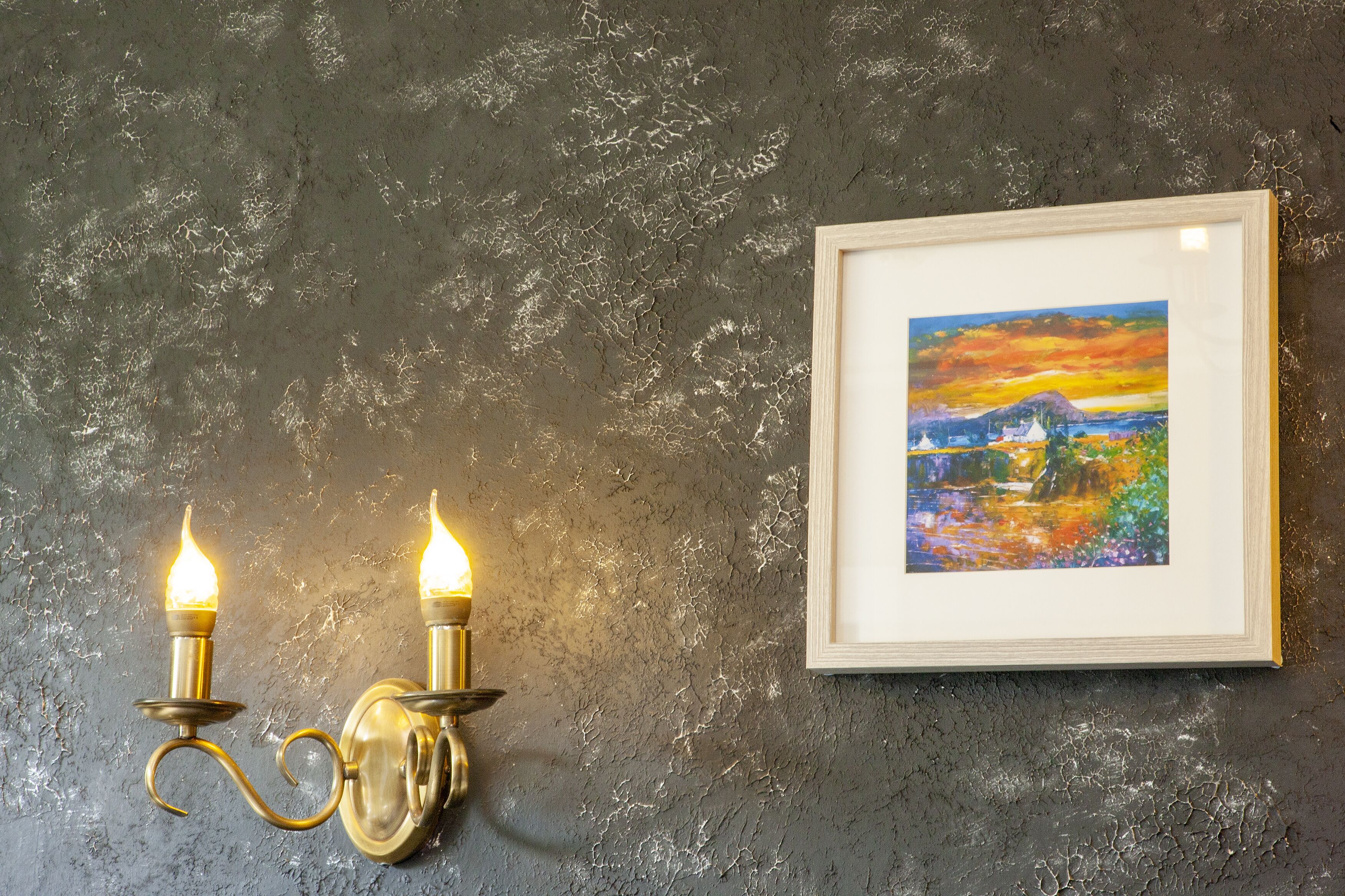 A textured gray wall with a framed painting of a landscape and two wall-mounted candelabra.