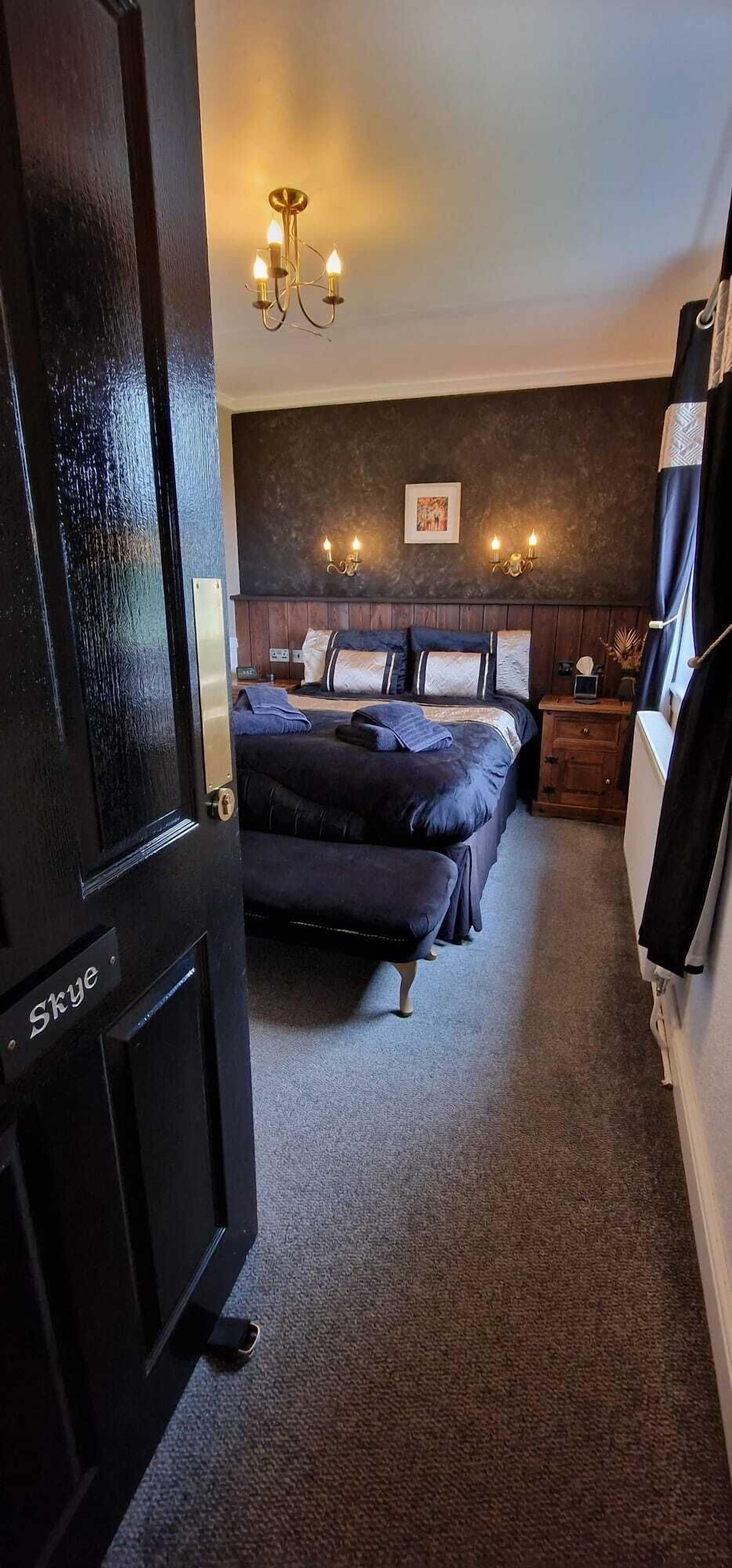 A bedroom with a dark wood door labeled "Skye," a large bed with dark bedding, a bedside table with a lamp, and a wall-mounted light fixture.
