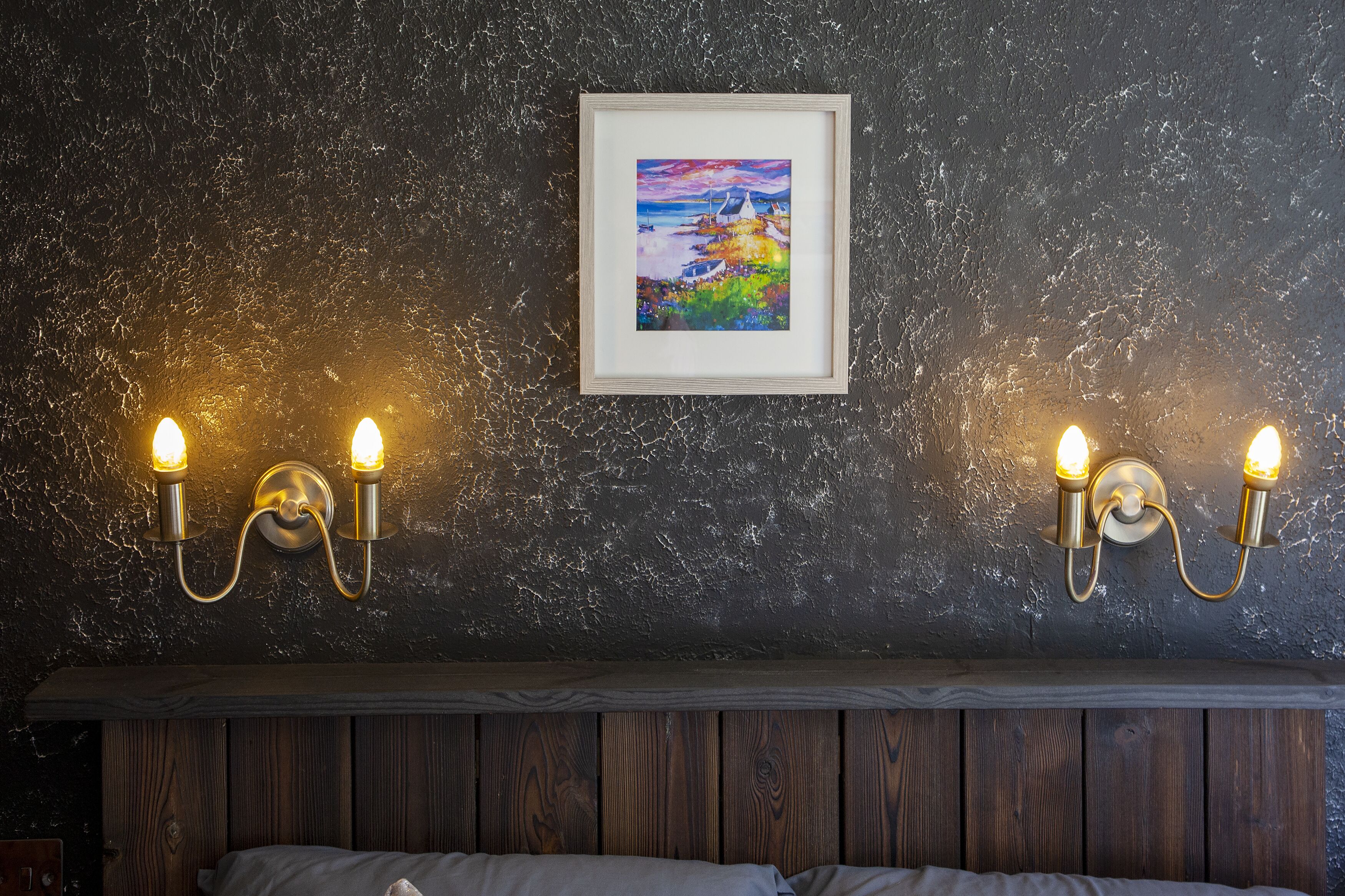 A room with a dark textured wall, a framed painting, and two wall-mounted light fixtures.