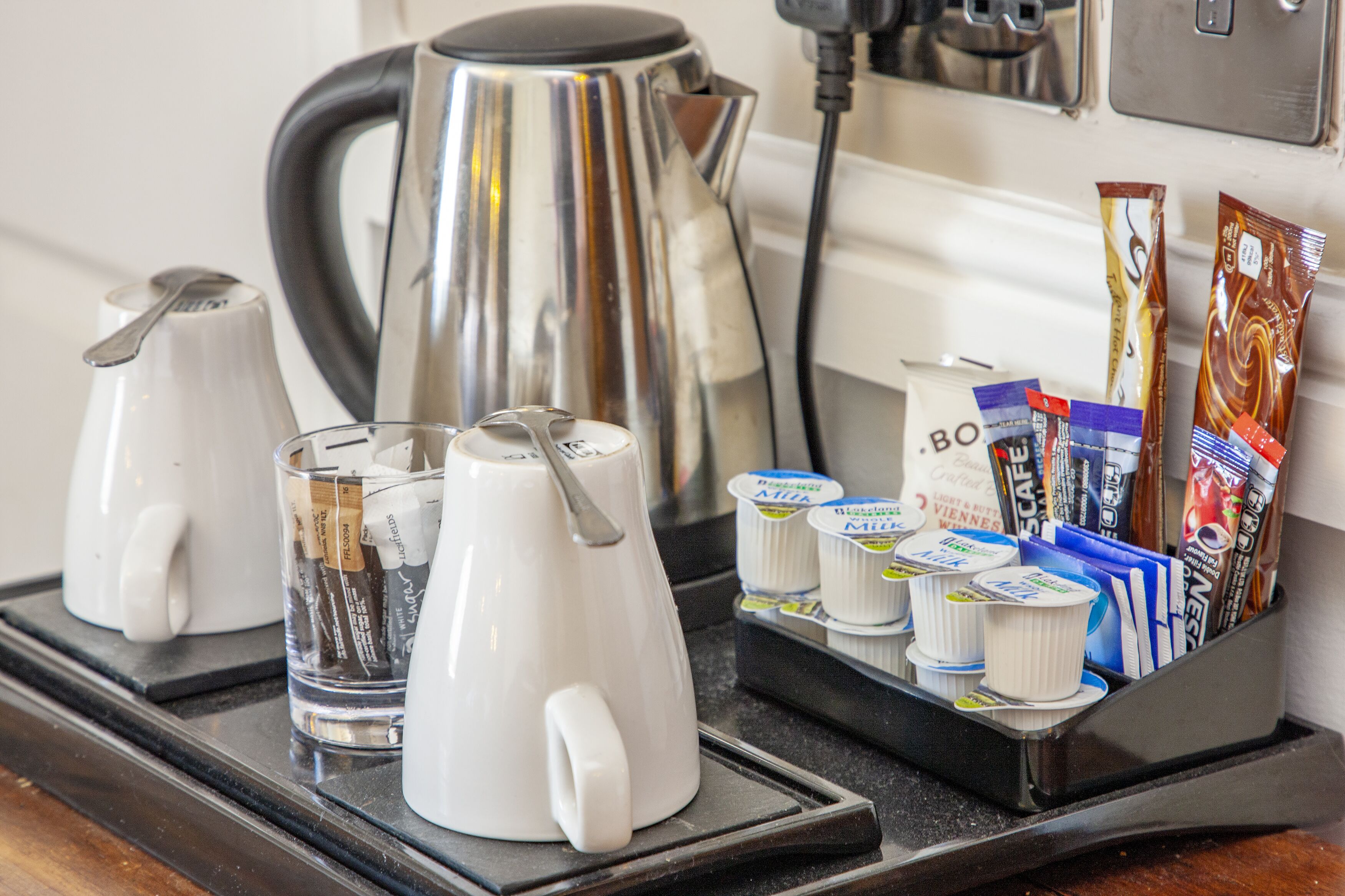 Una estación de café con una cafetera, dos tazas de café y una bandeja con cápsulas de café y sobres de crema.