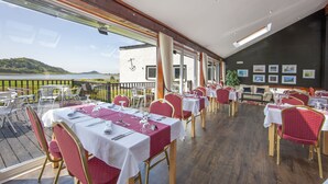 A restaurant with tables set for dining, a view of the sea, and a building with a white facade and anchor decoration. - The Galley of Lorne Inn (Lochgilphead)