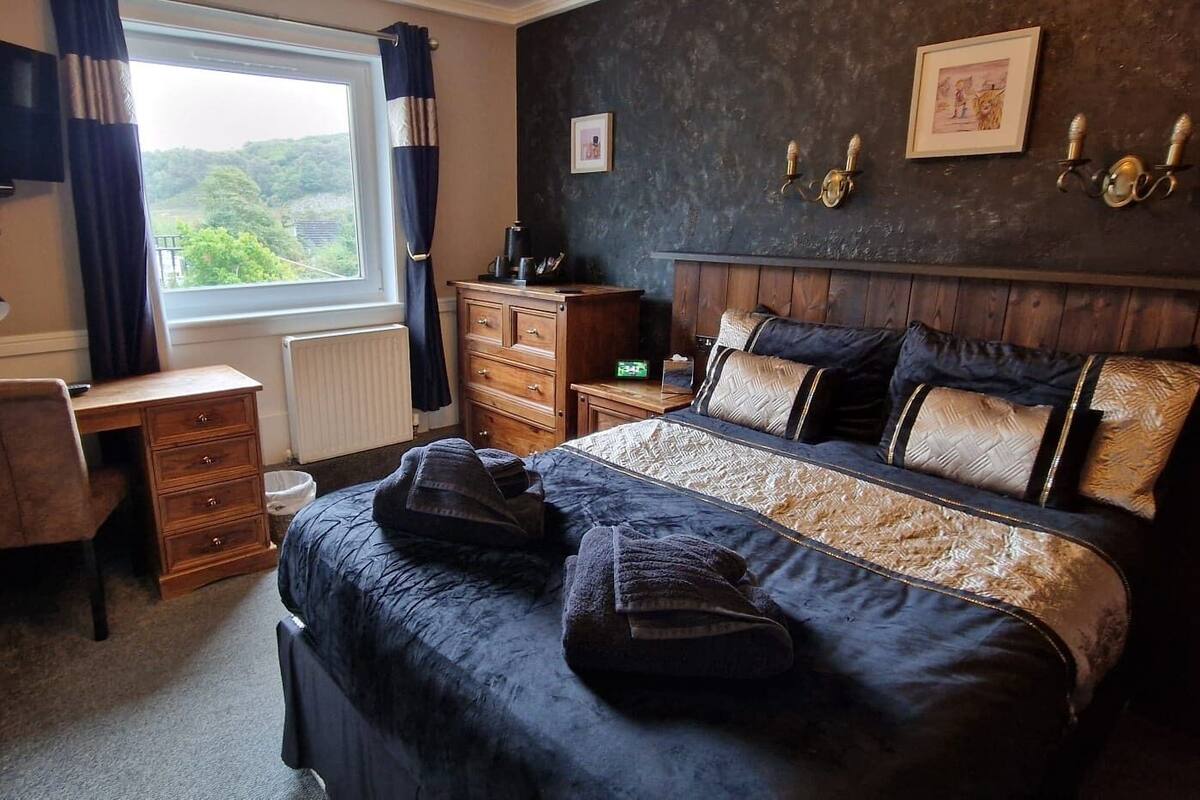 Un dormitorio con cama, escritorio y cómoda. Hay una ventana con cortinas, un televisor montado en la pared y un cuadro enmarcado.