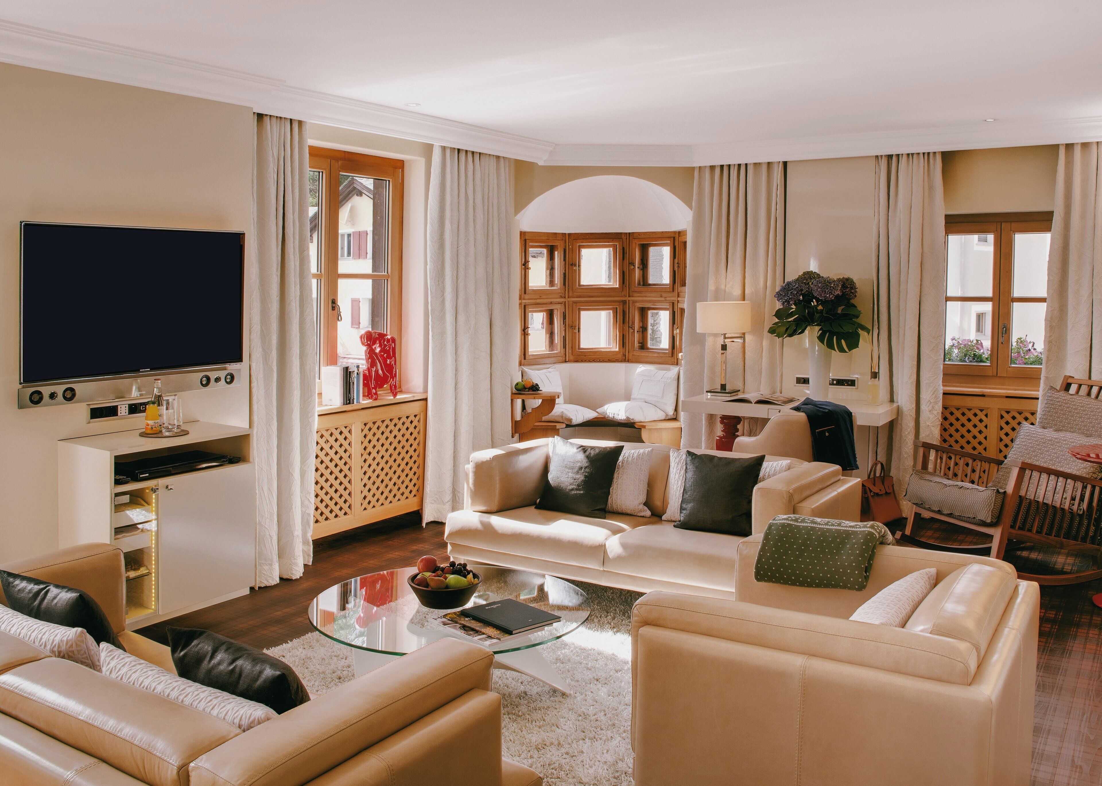 A well-lit living room with a flat-screen TV, leather sofas, a glass coffee table, and a view of a building through large windows.