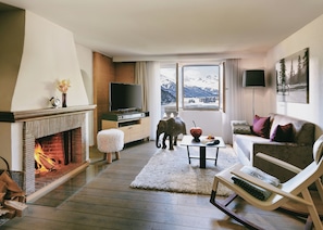 Ein gemütliches Wohnzimmer mit Kamin, Fernseher, Sofa und Blick auf die Berge durch das Fenster.