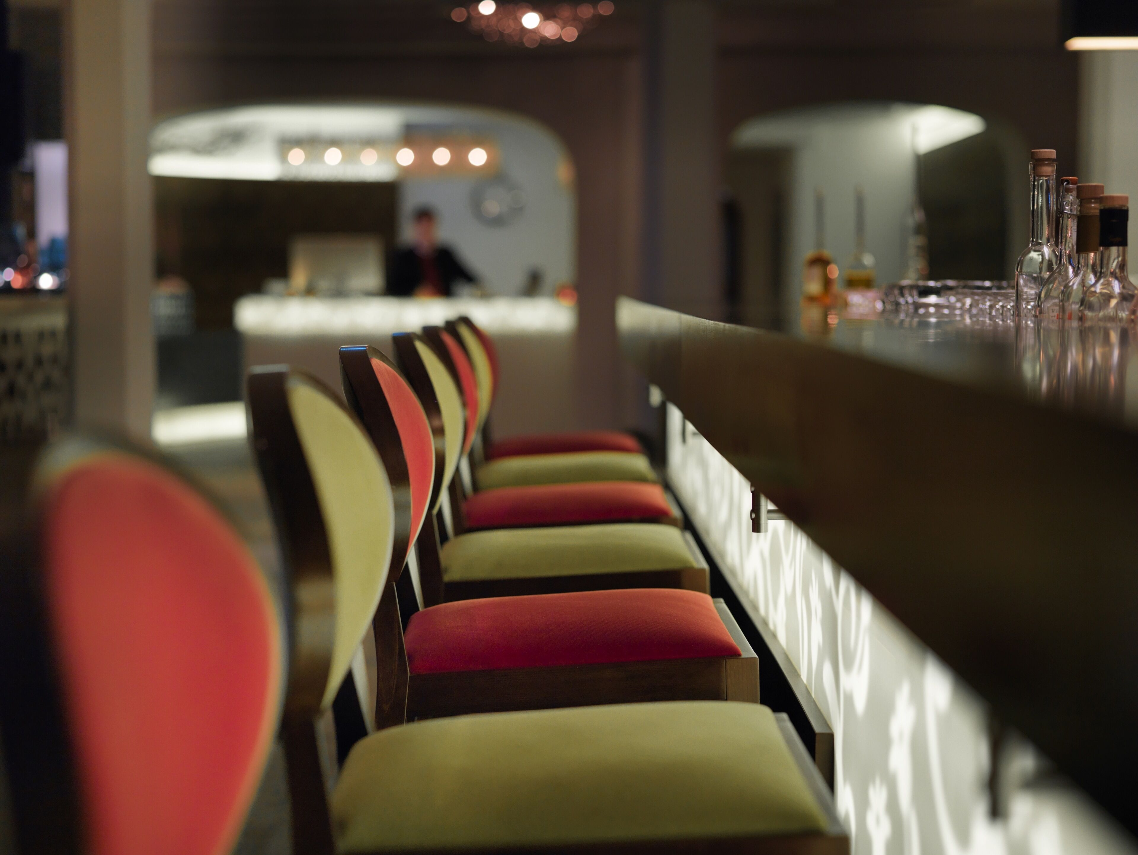 A row of chairs with cushioned backs and armrests, a bar area with bottles, and a person in the background.