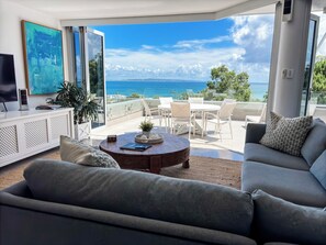 A modern living room with a large window, a sofa, a coffee table, and a TV. - Picture Point Terraces (Noosa Heads)