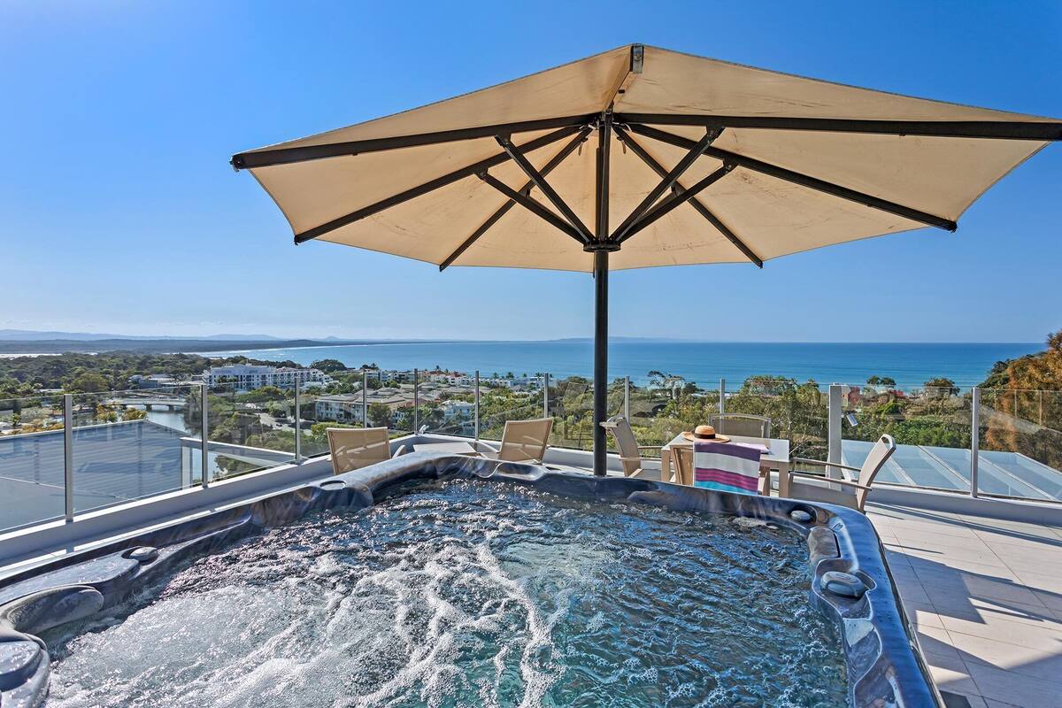 A hot tub with a view of the ocean, an umbrella, and a glass balustrade.