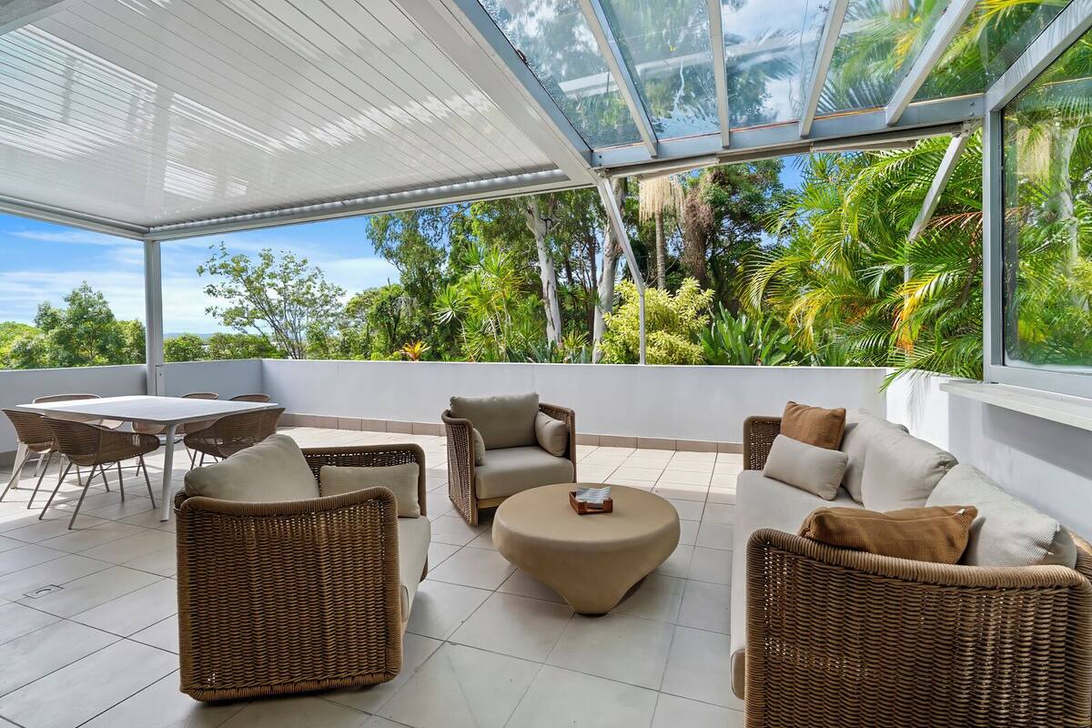 A covered outdoor area with wicker furniture, a round table, and a view of lush greenery.