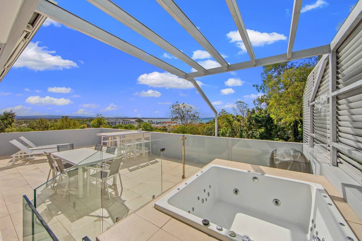 A rooftop terrace with a glass-enclosed hot tub, lounge chairs, and a clear view of the ocean and cityscape.