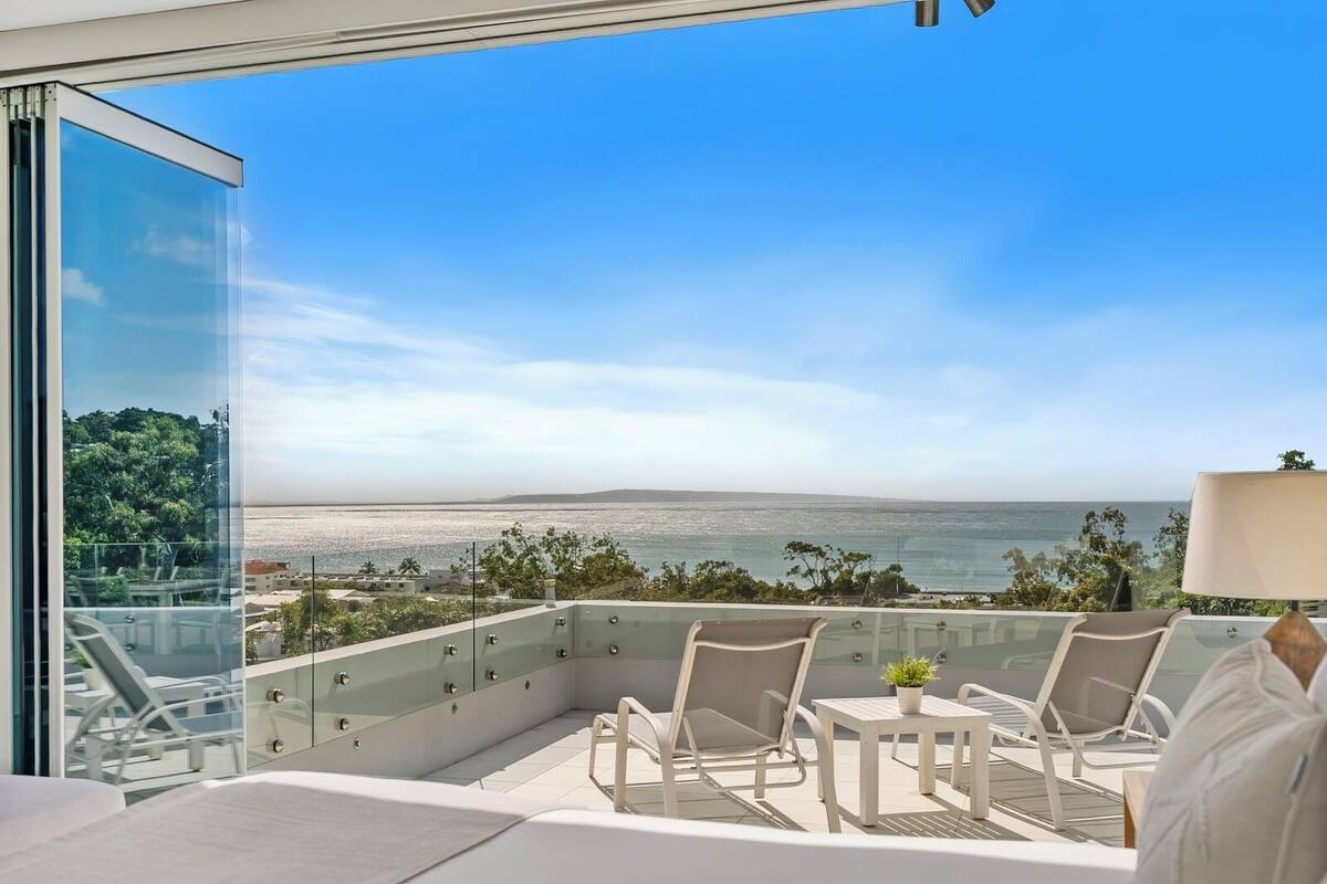 A balcony with a glass railing, a table, and two chairs, overlooking a coastal view.