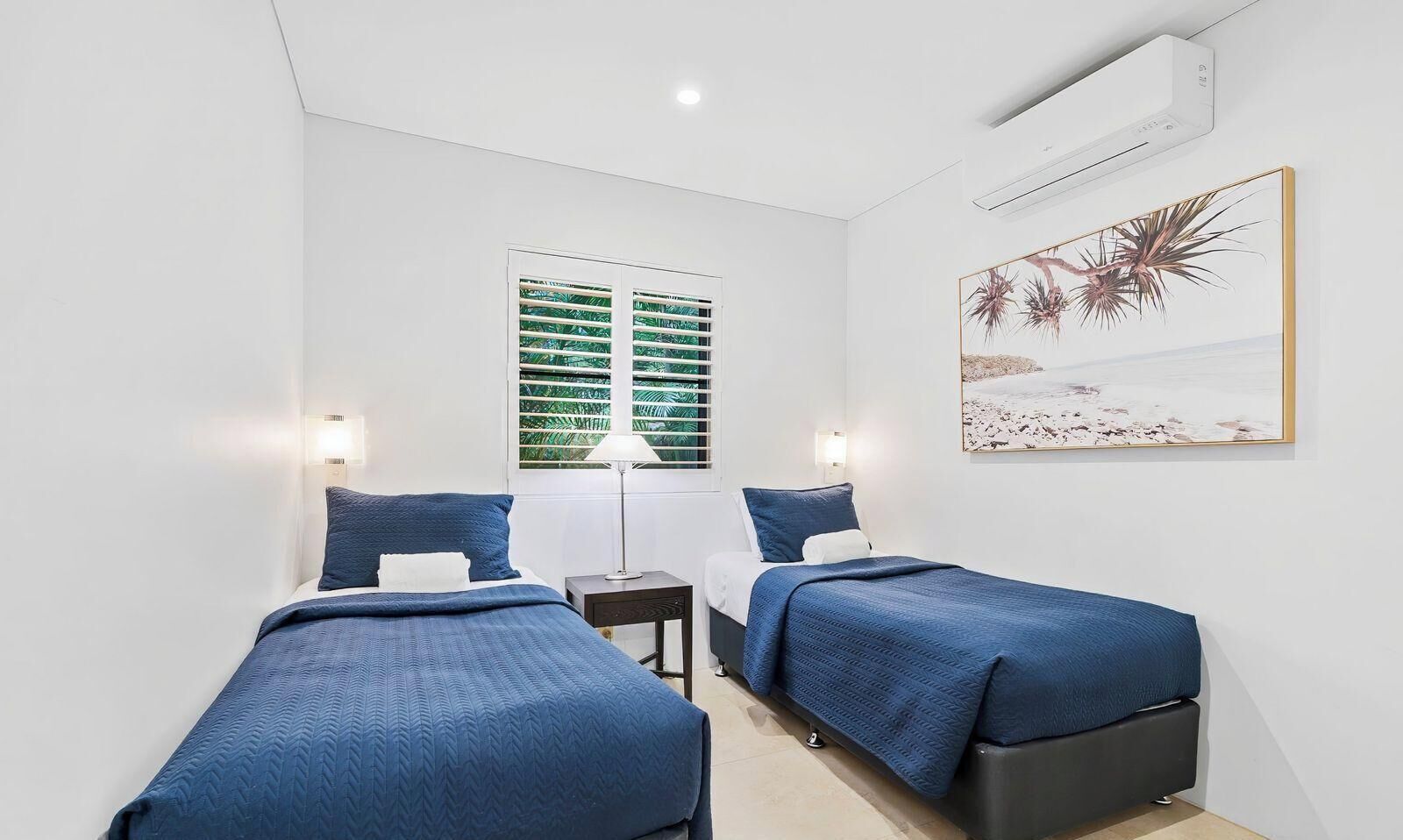 A room with two beds, a window with blinds, a wall-mounted air conditioner, and a framed picture.