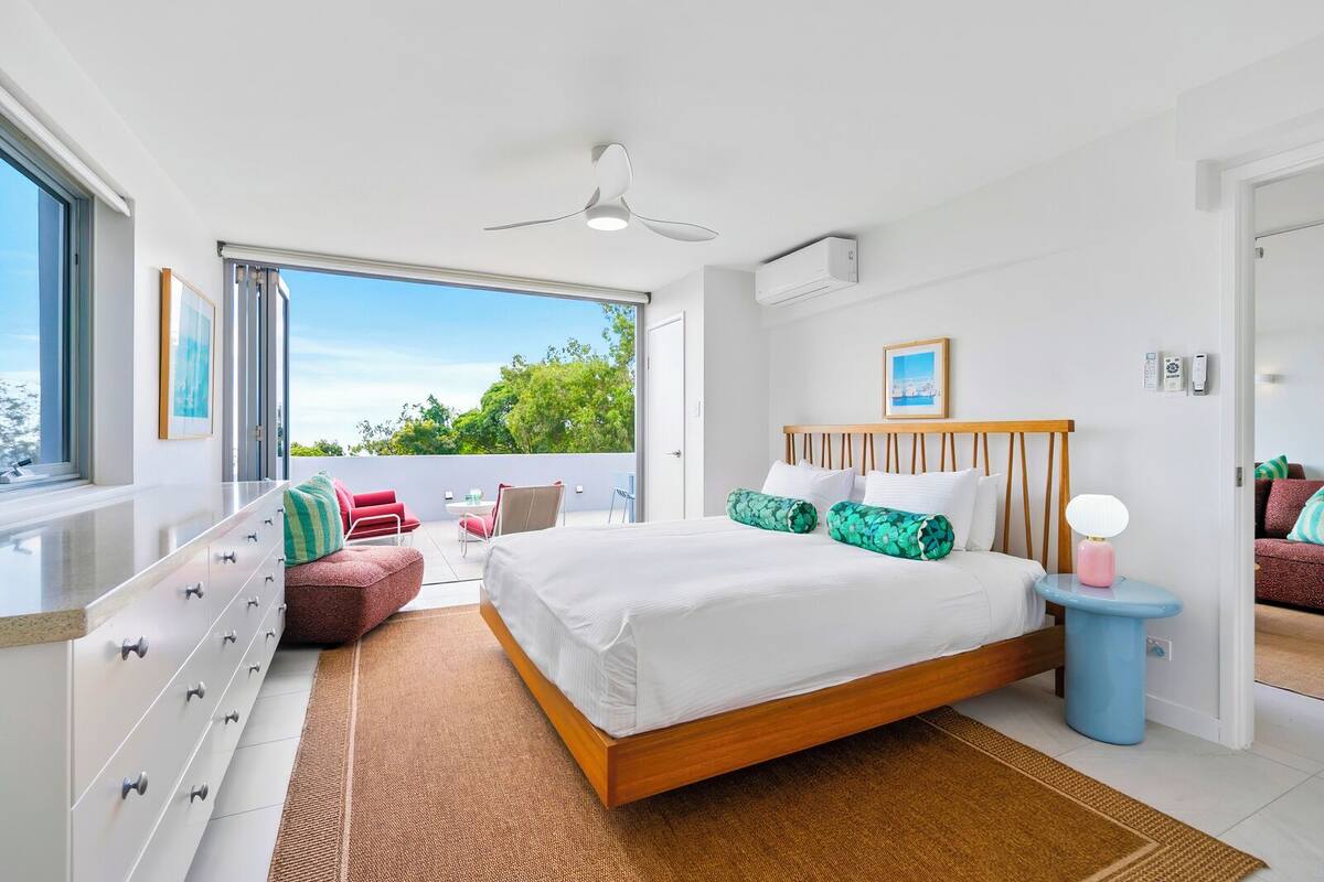 A modern bedroom with a large bed, a ceiling fan, a view to the outdoors, and a balcony with seating.