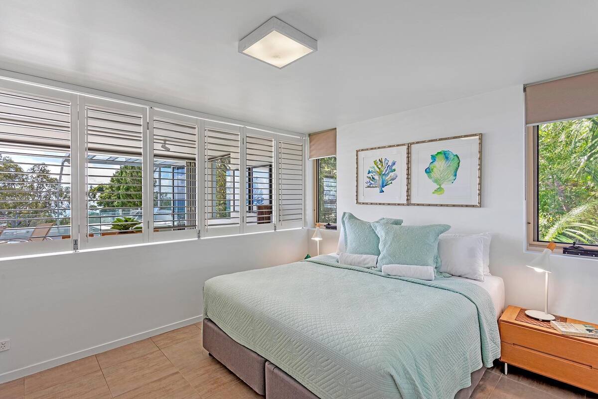 A bedroom with a bed, a nightstand, a lamp, a window with blinds, and two framed artworks on the wall.