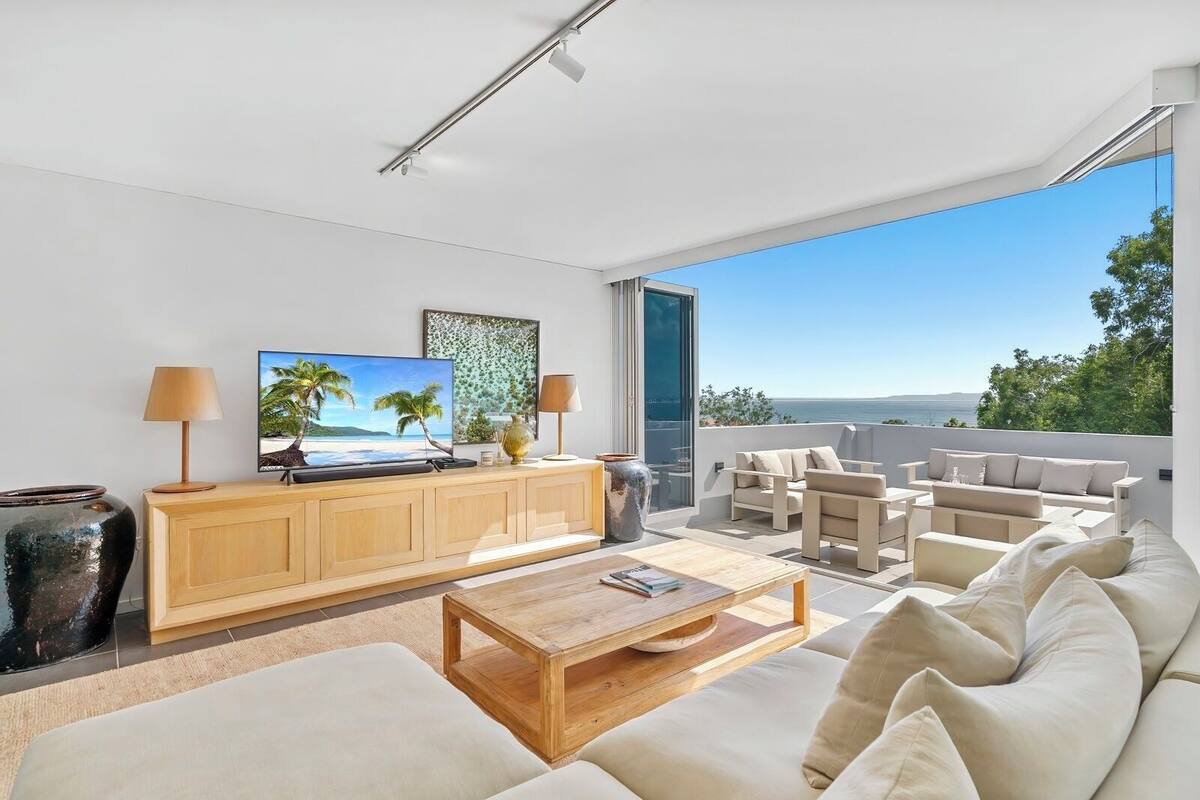 A modern living room with a flat-screen TV, wooden furniture, a large window with a view of the sea, and a balcony with outdoor seating.