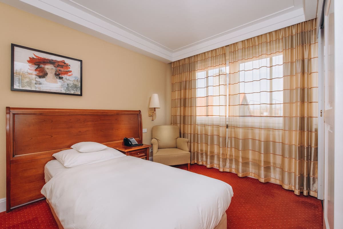 A hotel room with a large bed, a wooden headboard, a chair, a bedside table with a lamp, and a window with curtains.