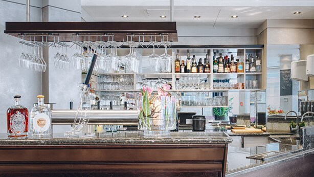 Uma área de bar com bancada de granito polido, copos de vinho pendurados e prateleiras repletas de garrafas de bebidas diversas.