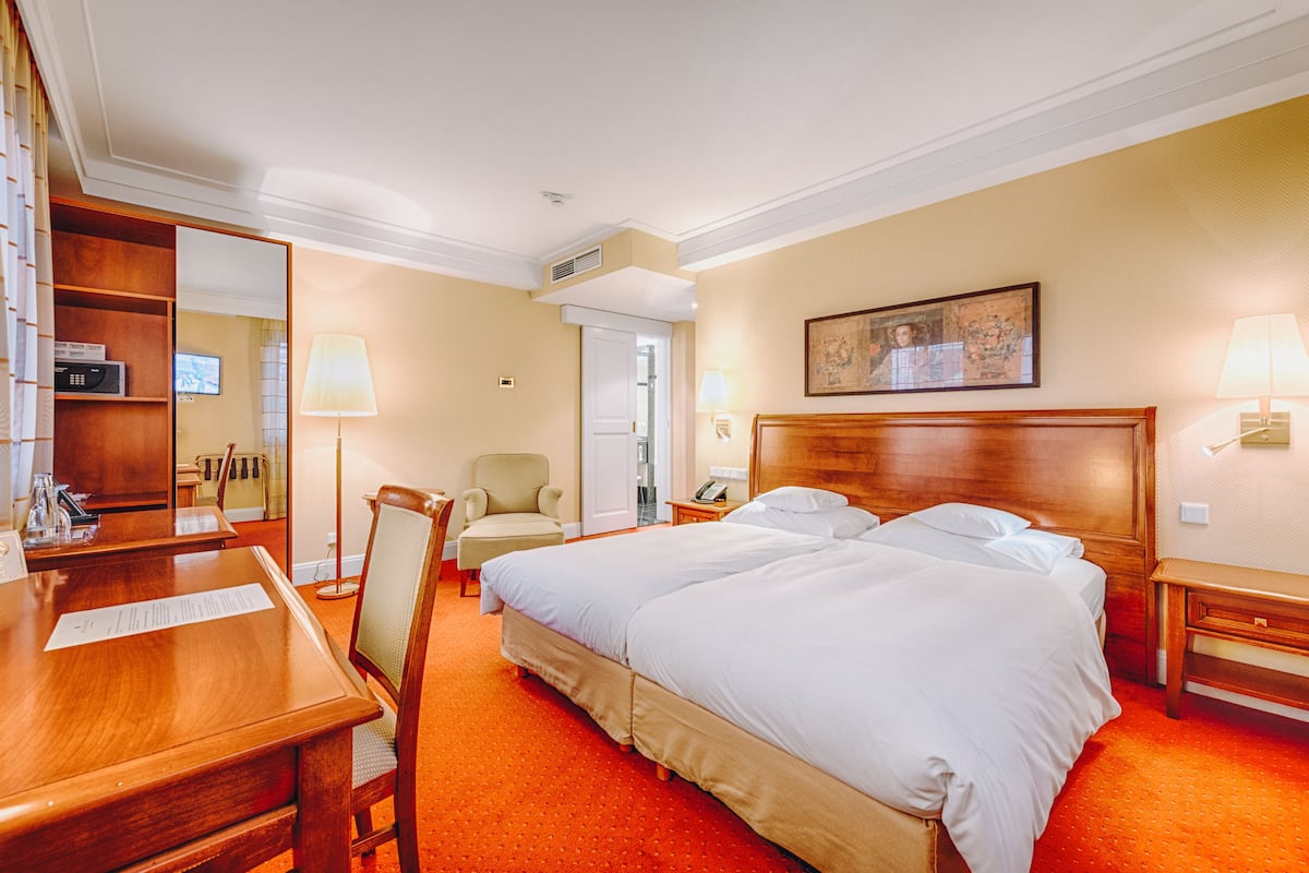 A hotel room with a large bed, a desk, a chair, a bedside table, a lamp, and a painting on the wall.