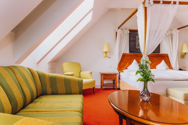 Una habitación de hotel con cama, sofá, mesita de noche, lámpara y un florero con flores.