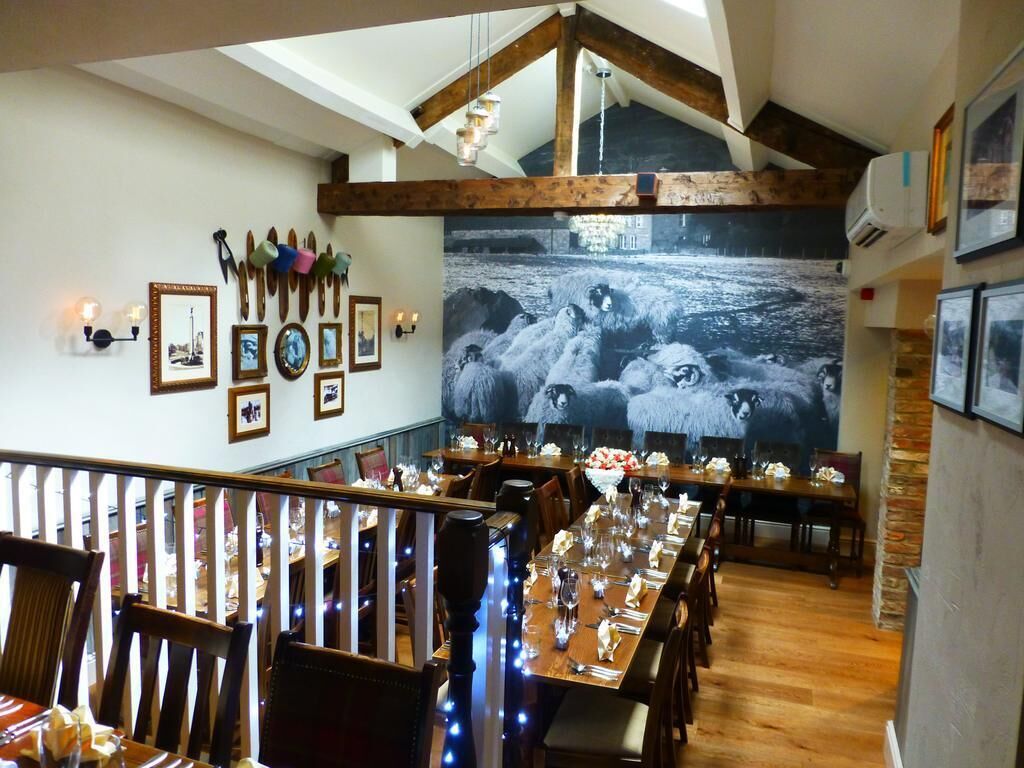 Un coin repas avec des tables et des chaises en bois, un grand tableau en noir et blanc représentant des moutons accroché au mur, et un escalier muni d’une rampe.
