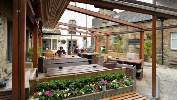 An outdoor patio area with wooden benches and tables, stone walls, and hanging flower baskets. - The Woolly Sheep Inn (Skipton)