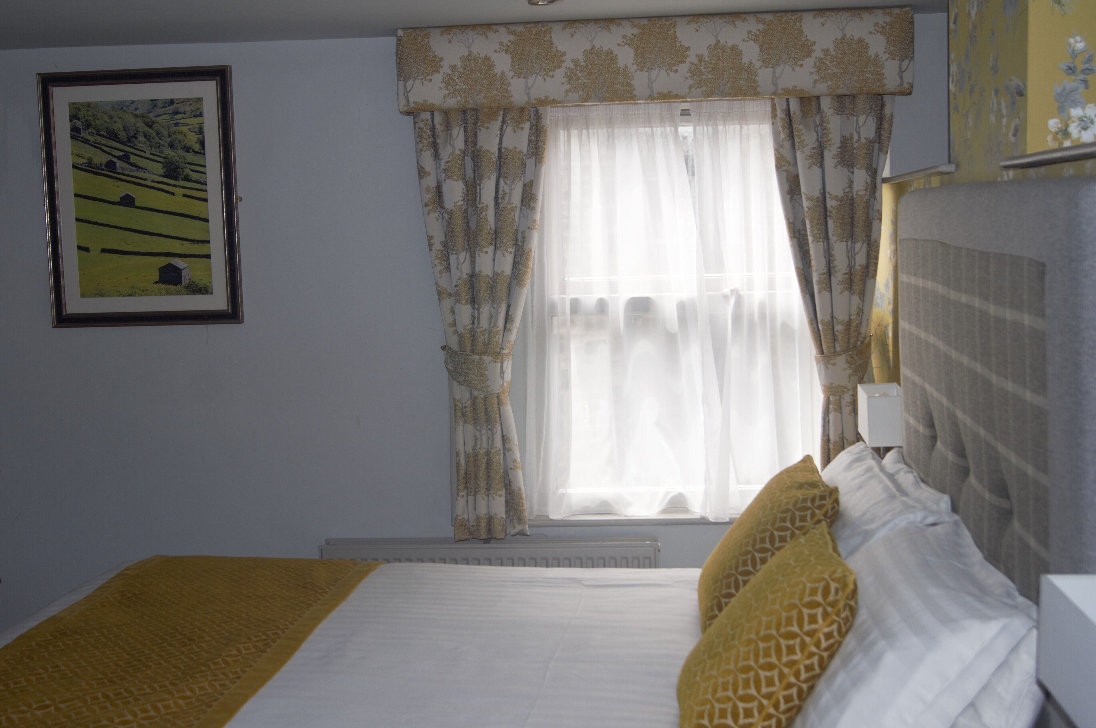 Un dormitorio con cama, ventana con cortinas, radiador y un cuadro en la pared.