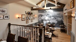 A dining area with wooden tables and chairs, a staircase with a white railing, and a large black and white photograph on the wall. - The Woolly Sheep Inn (Skipton)