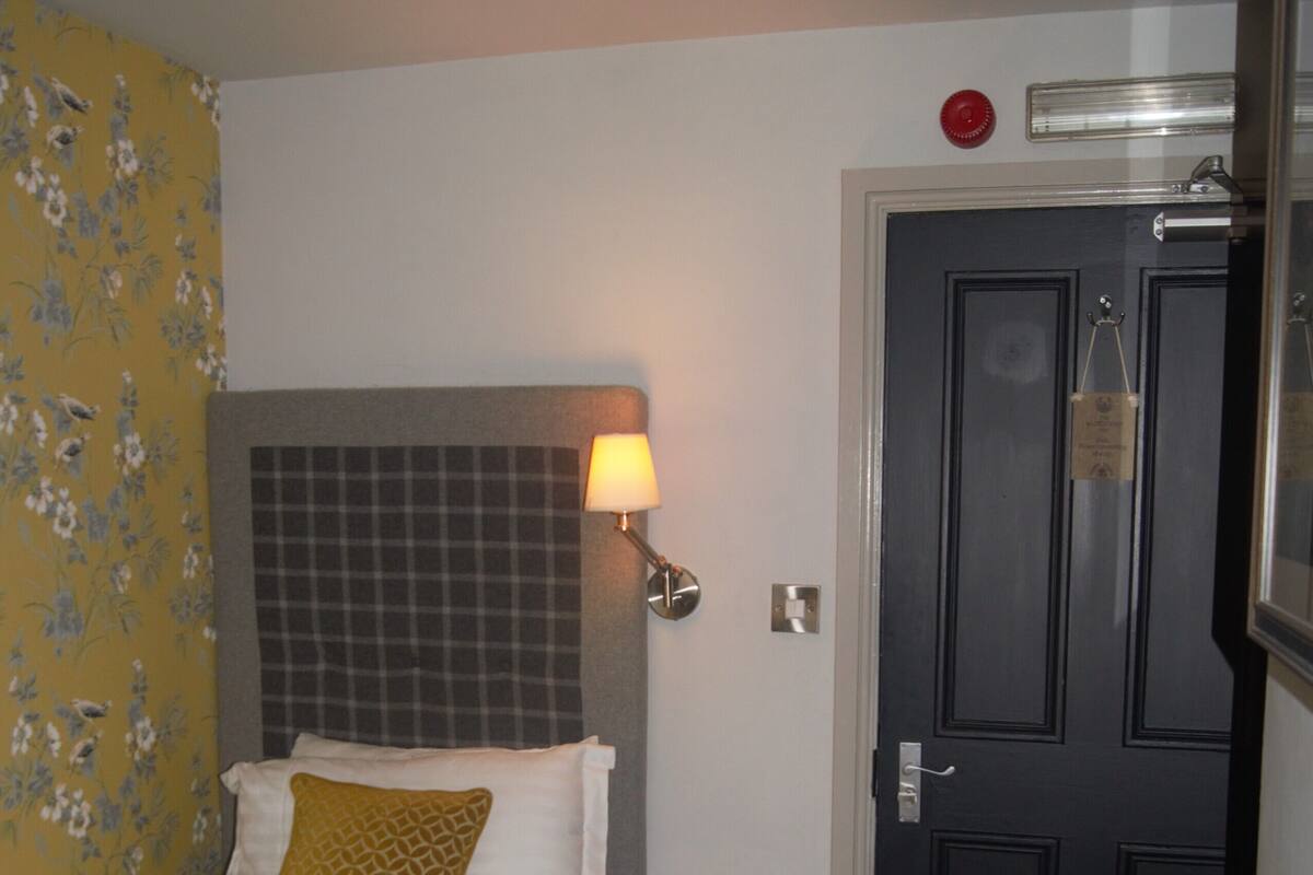 A hotel room with a bed, a nightstand, a lamp, a door, and a wall with floral wallpaper.