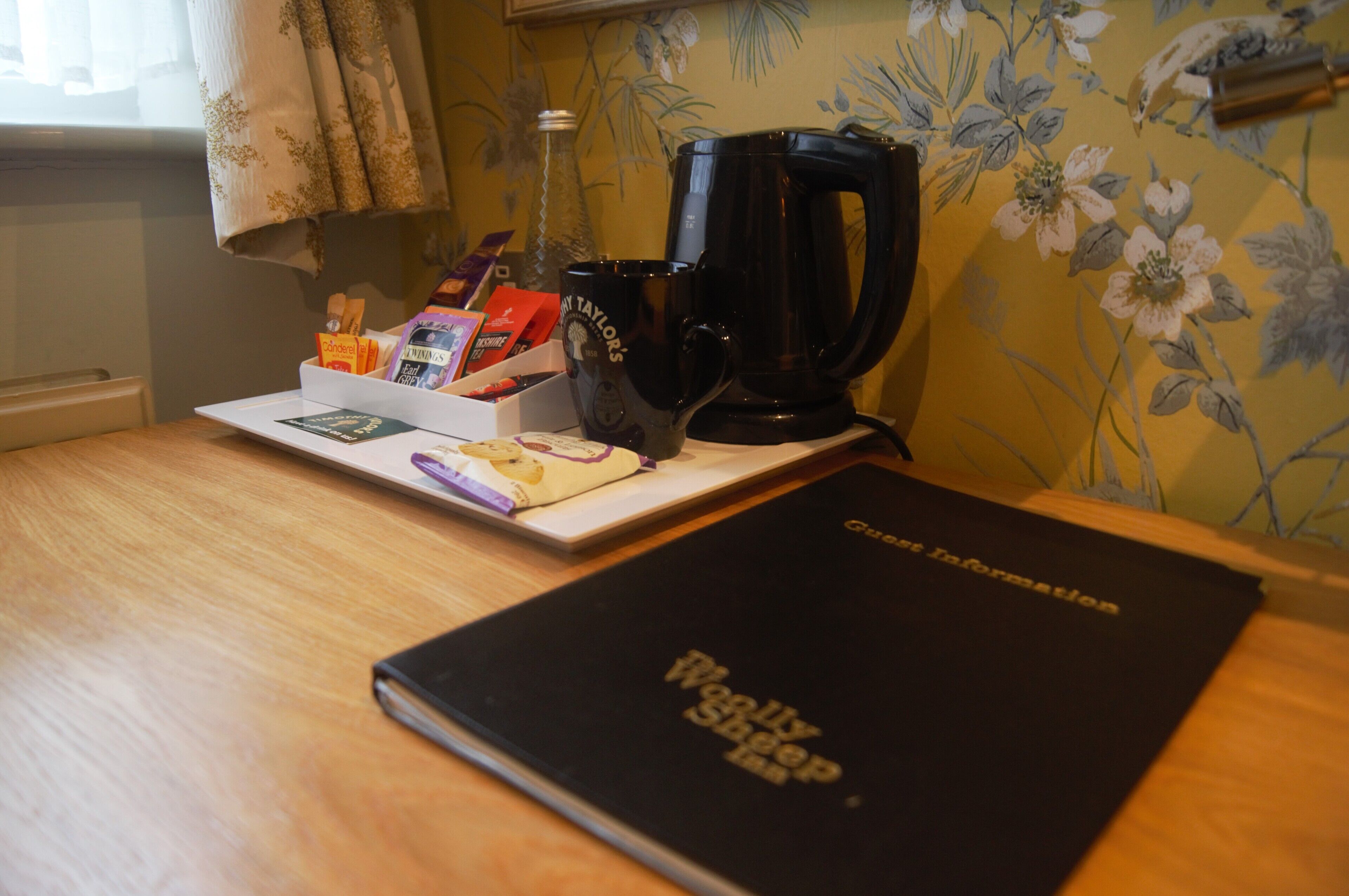 A wooden table with a black notebook, a black coffee maker, and a tray with various items including a purple pen and a small box.
