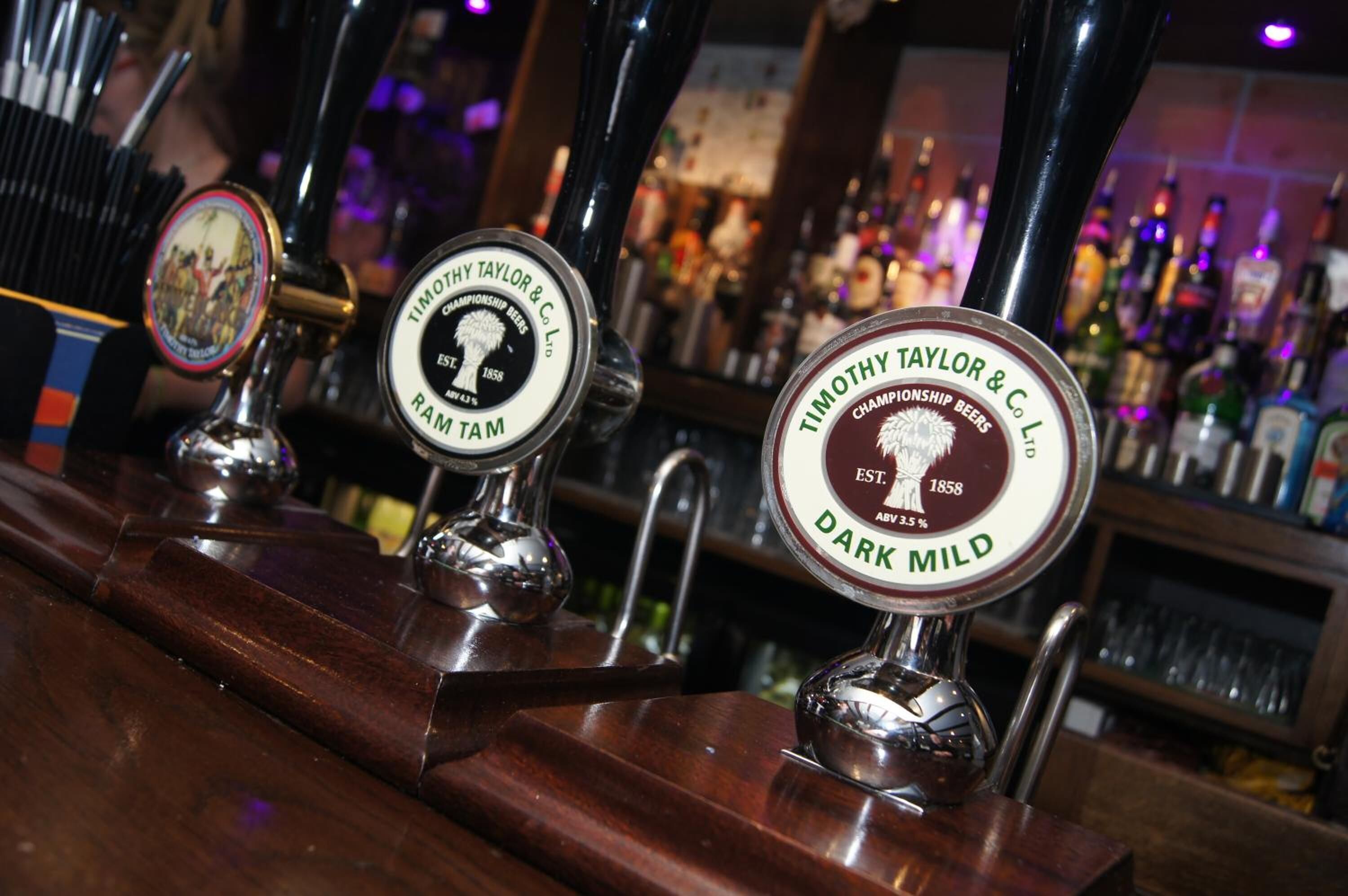 Un bar con varias llaves de cerveza que indican 'Timothy Taylor & Co. Cervezas Campeonas' y 'Dark Mild'.