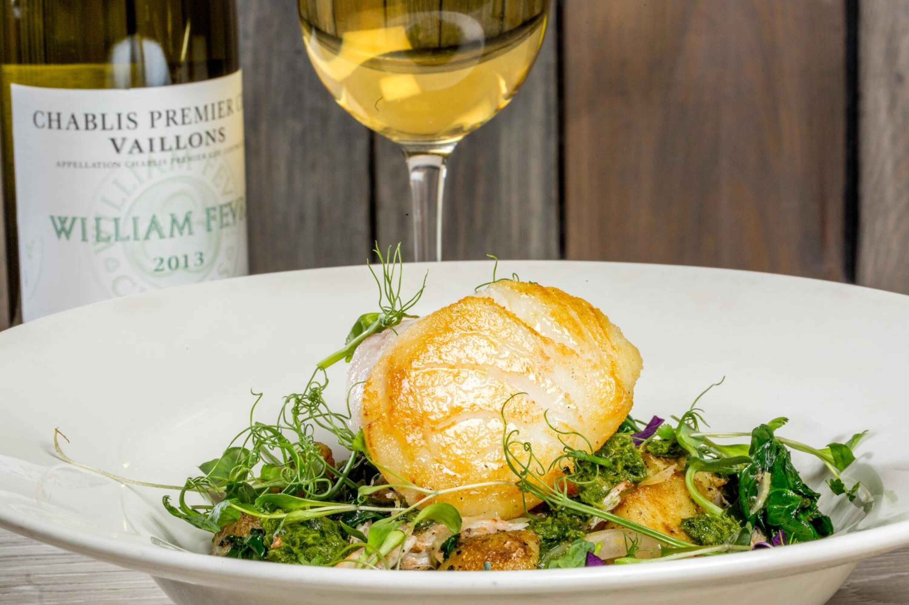 Une assiette de nourriture comprenant une noix de Saint-Jacques et des légumes verts, avec une bouteille de vin en arrière-plan.