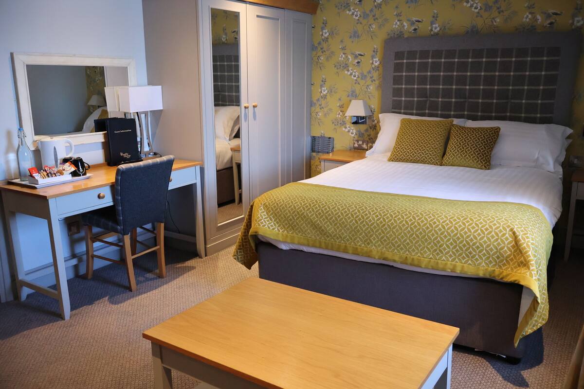 A hotel room with a bed, a desk with a chair, a mirror, and a bedside table.
