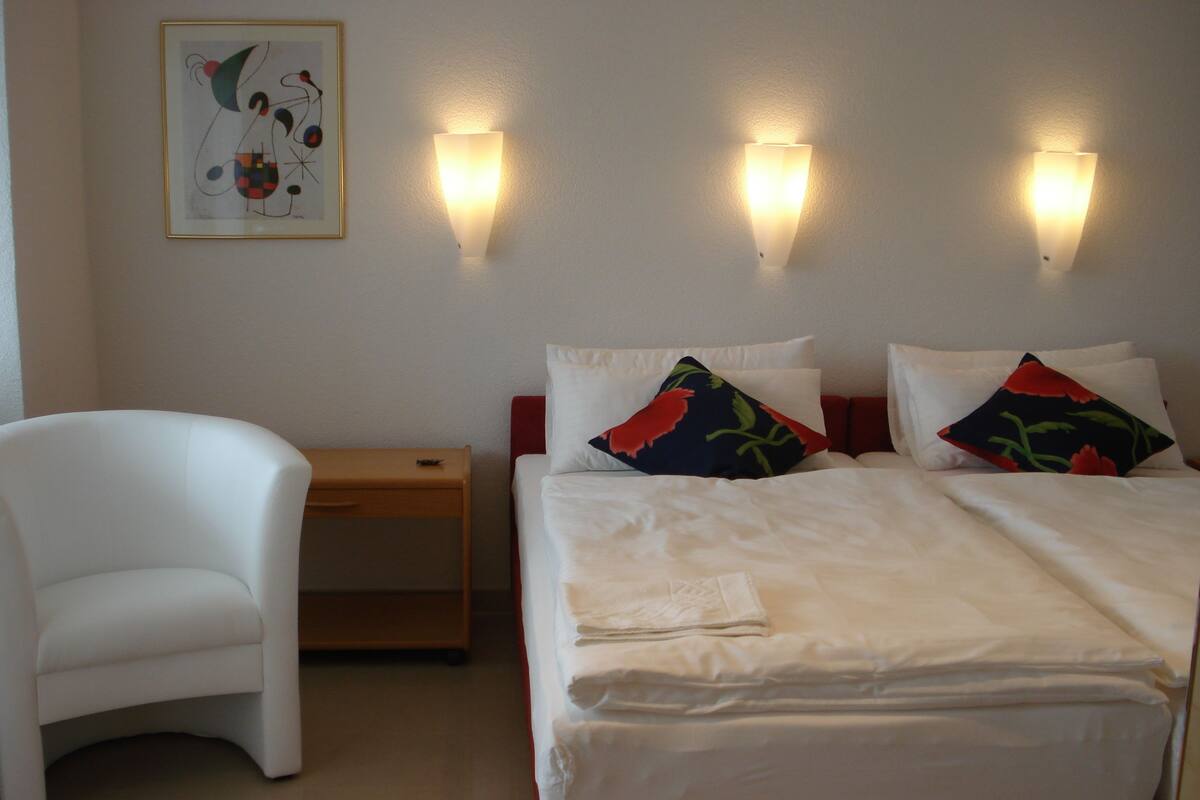 A hotel room with a bed, two pillows, a chair, a nightstand, and a wall-mounted light fixture.