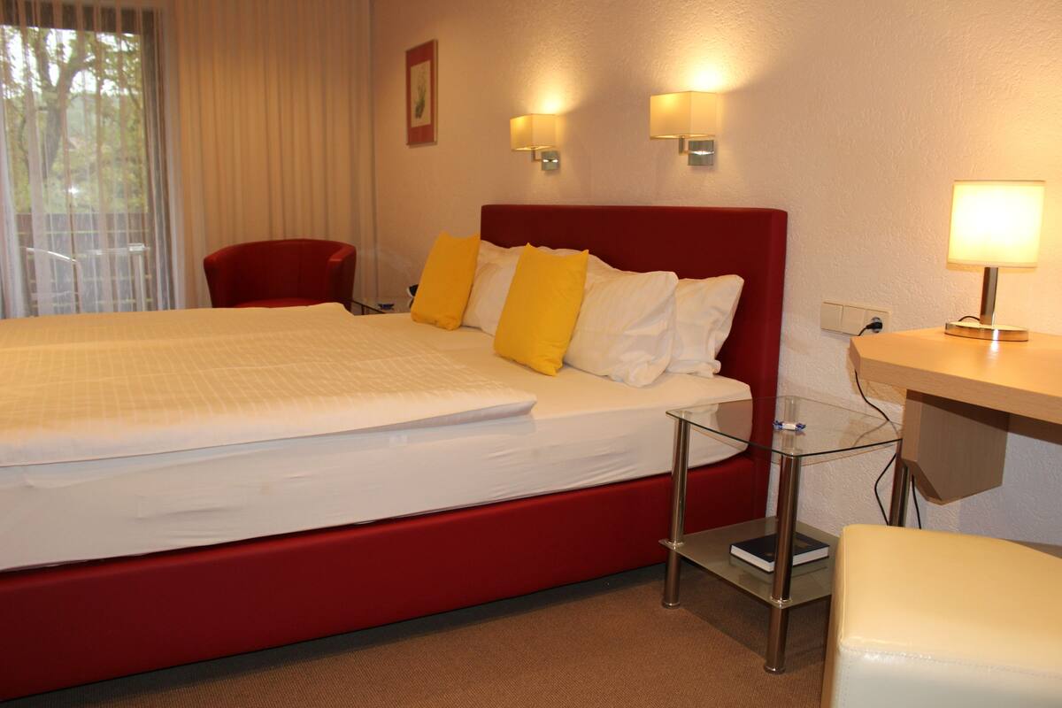 A hotel room with a large bed, a red headboard, two bedside lamps, a glass side table, and a red chair.
